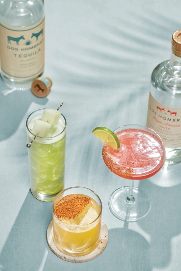 All Cocktails Together Fantasy Land News Green mojito with ice and melon cubes pink margarita with lime wedge on a light blue table tequila bottles visible in the background