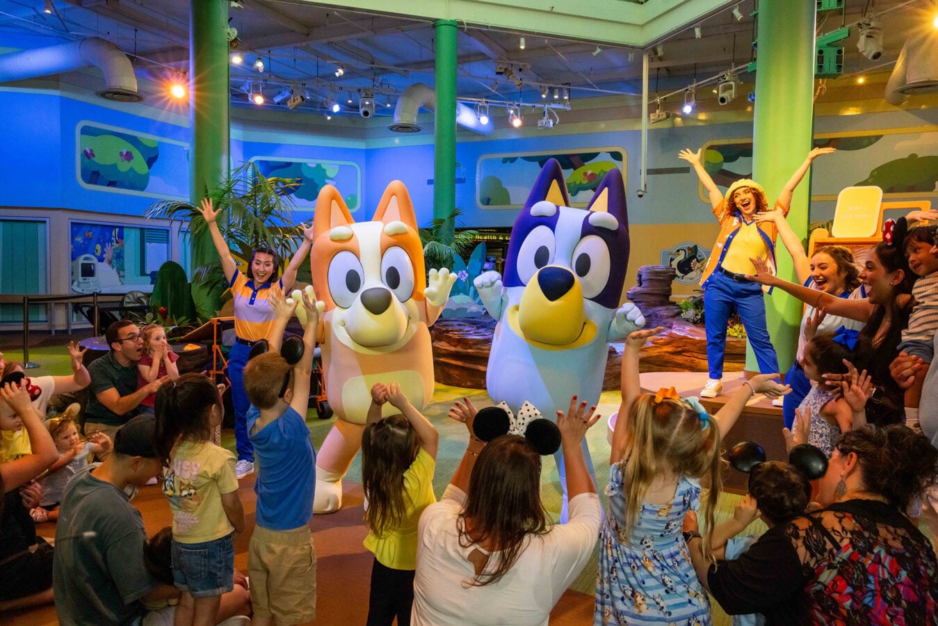 Children cheer and reach toward two large colorful cartoon dog mascots on a stage in a bright indoor show for kids