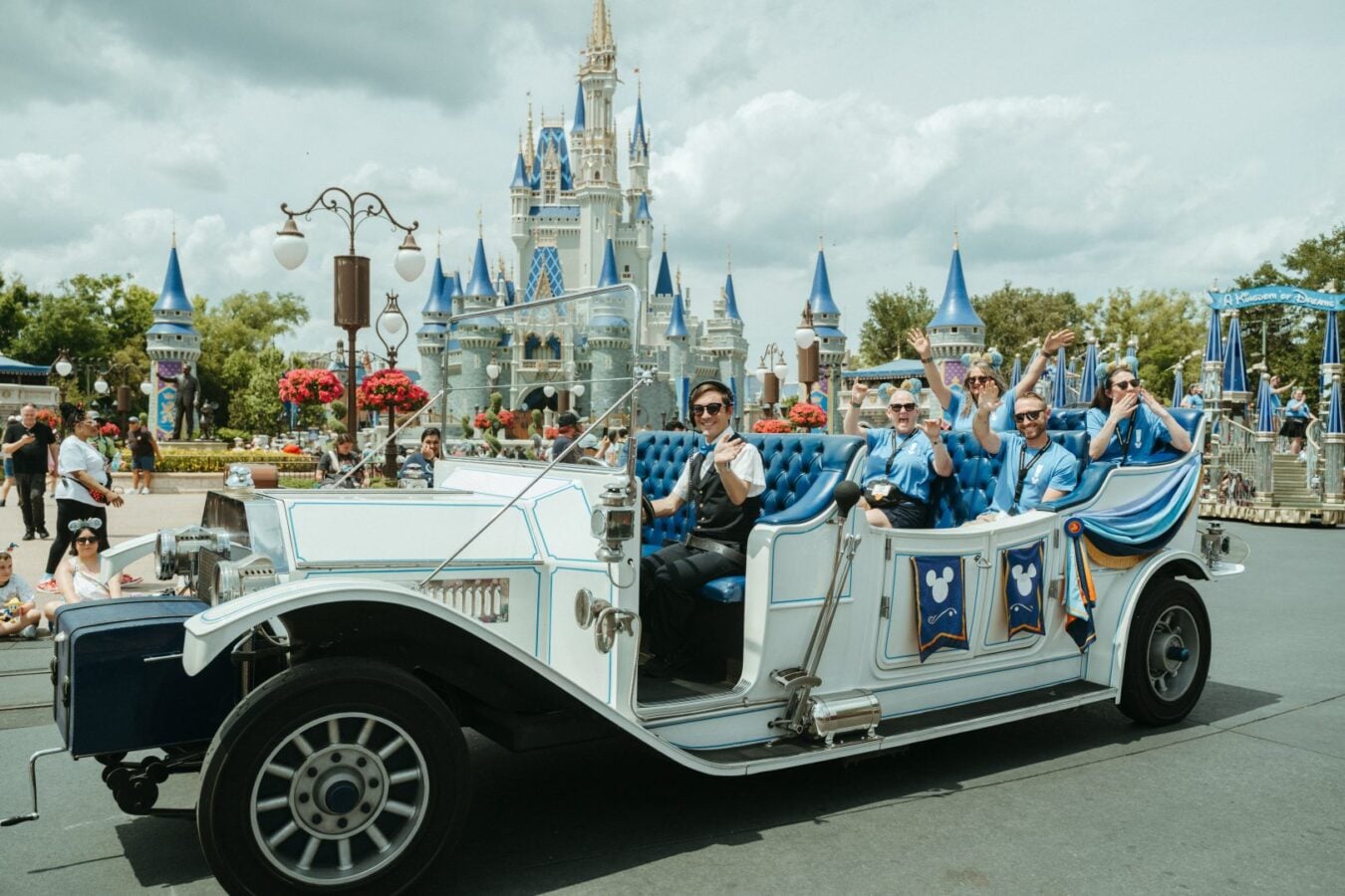 Vintage Disney parade carriage with blue upholstery passes in front of Cinderella Castle riders wave to onlookers
