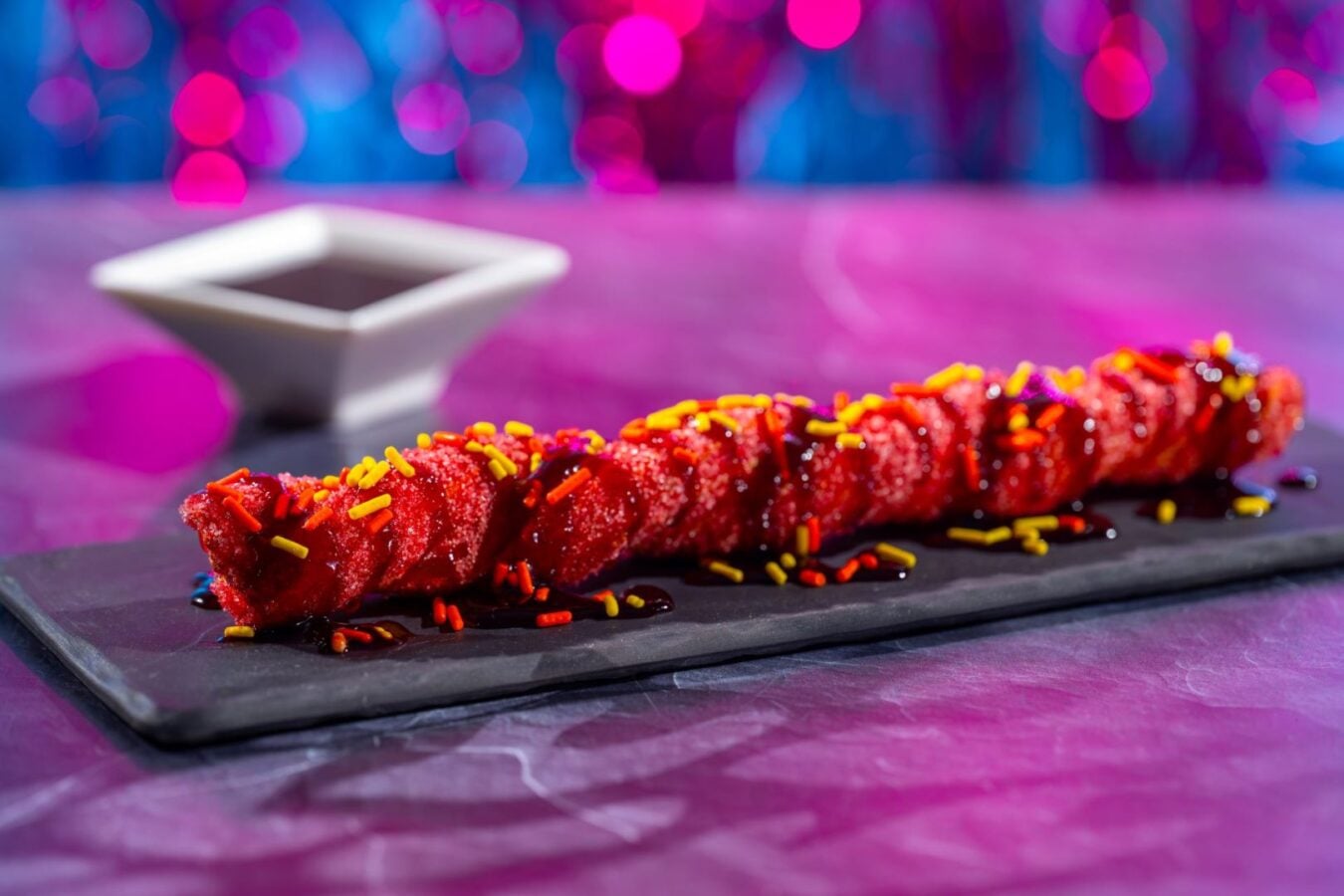 Red candied skewered dessert with yellow and orange sprinkles on a slate plate blurred white dipping bowl and neon purple pink background