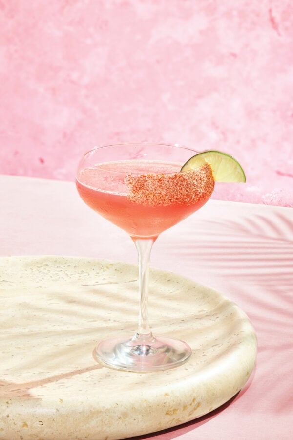 Cranstonian Fantasy Land News Pink cocktail in a sugar rimmed coupe glass with a lime wedge set on a light stone surface against a pink backdrop for a festive vibe