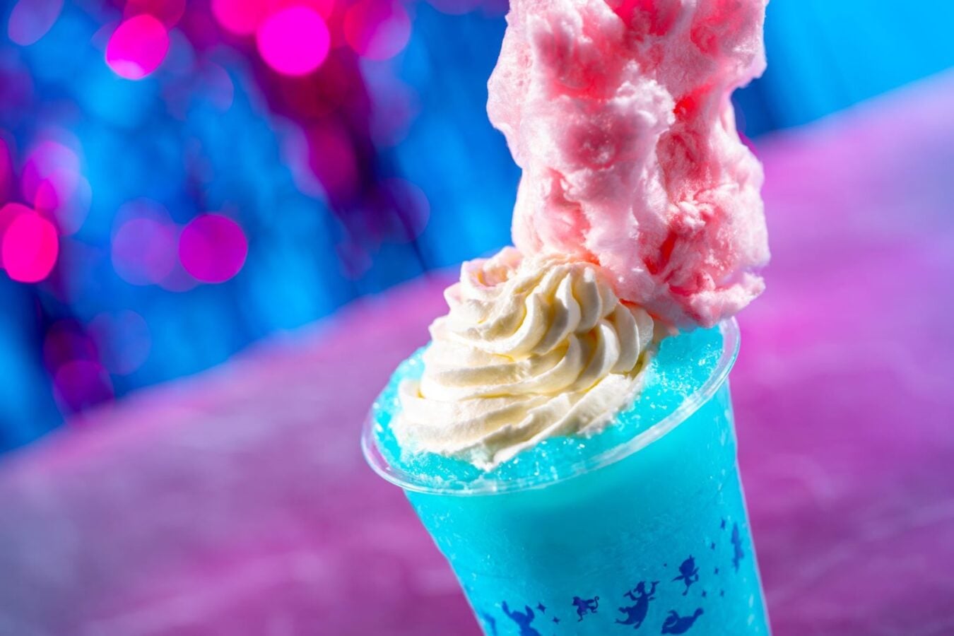 Blue slushy drink in a cup with whipped cream and pink cotton candy against a vibrant pink blue bokeh background