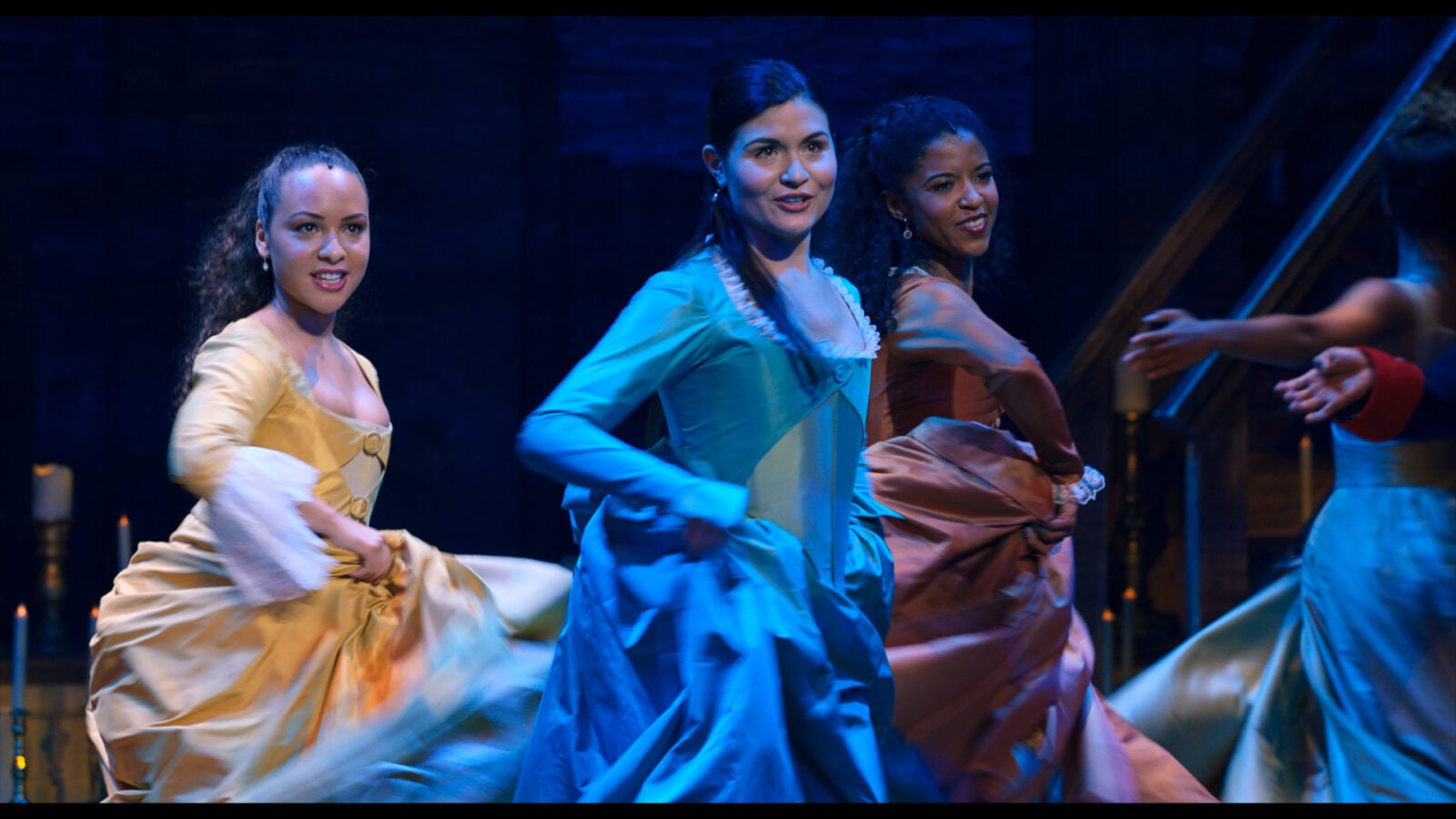 Three women in 18thcentury gowns perform a synchronized dance on a dimly lit stage with blue lighting