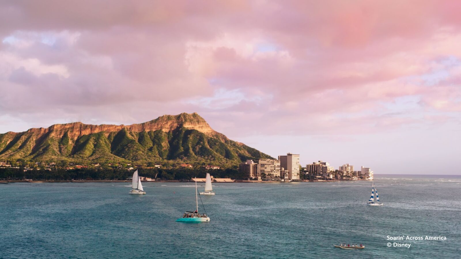 Sunset over a calm bay with sailboats a green ridged mountain Diamond Head in the distance and a city shoreline with buildings along the coast
