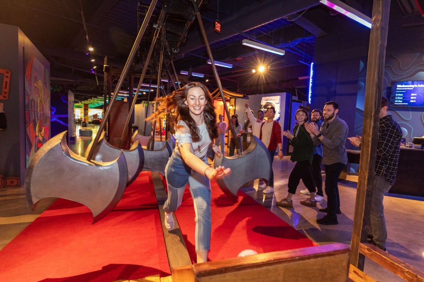 Woman in jeans runs toward the camera on a large bat winged ride while a cheering group watches in an arcade like venue