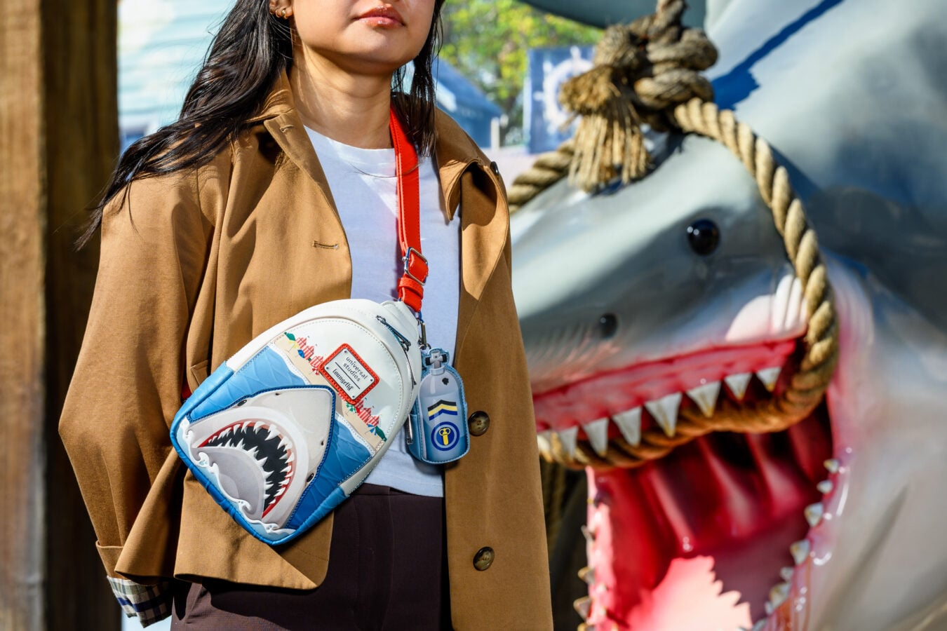 Person in a tan coat holds a blue shark graphic backpack with a red strap with a large dinosaur sculpture in the background