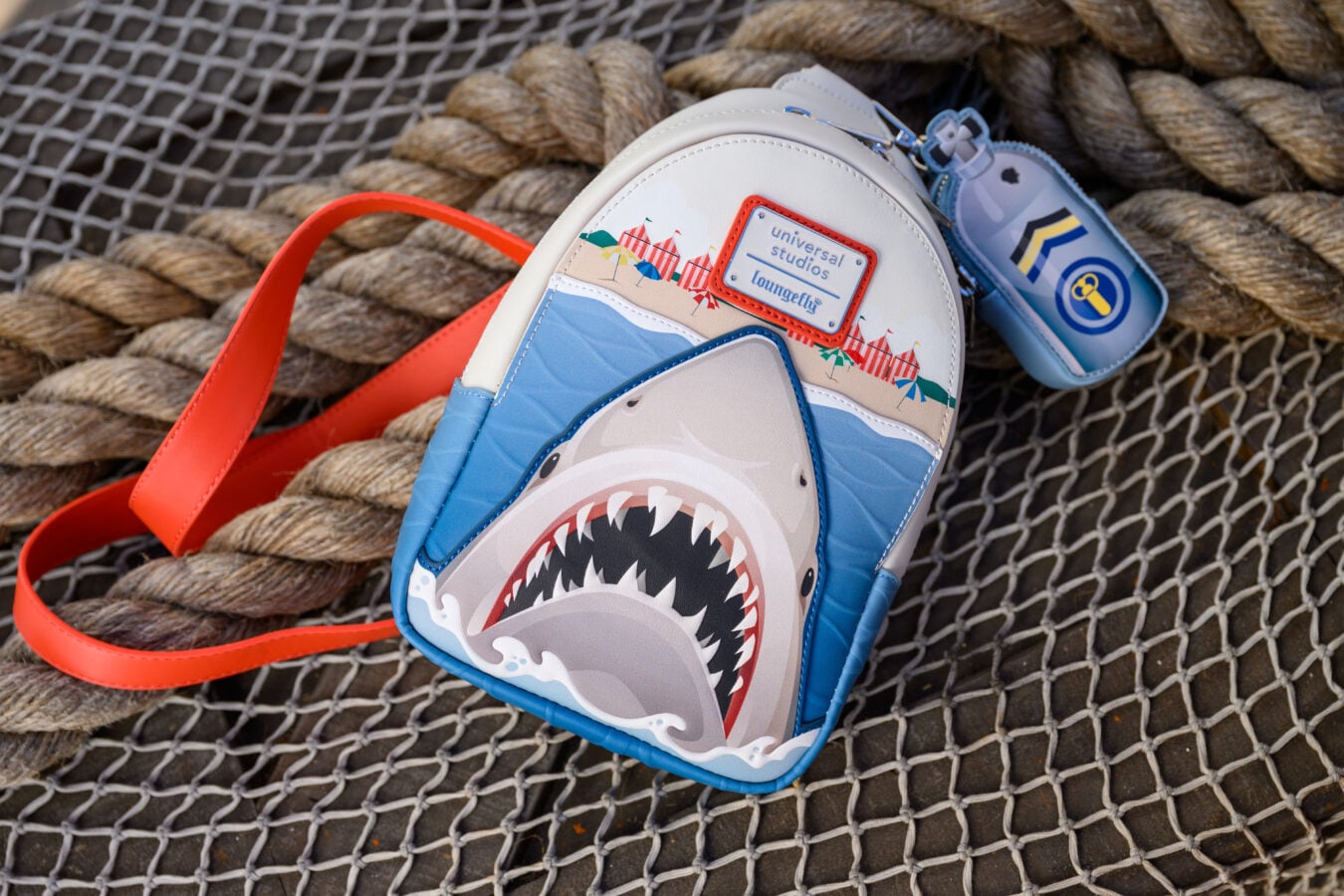 White and blue shark themed Loungefly backpack with a beach umbrellas graphic and red bordered tag on a rope net backdrop