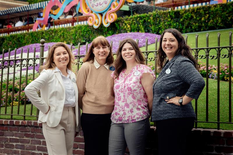 Four women smiling and posing together in front of a decorative iron fence at a colorful theme park garden behind them