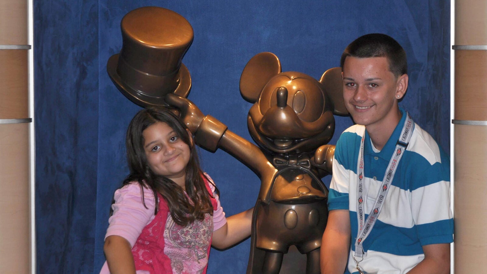 Two smiling children posing with a bronze Mickey Mouse statue in front of a blue backdrop hat tipped and bow tied both leaning toward the character