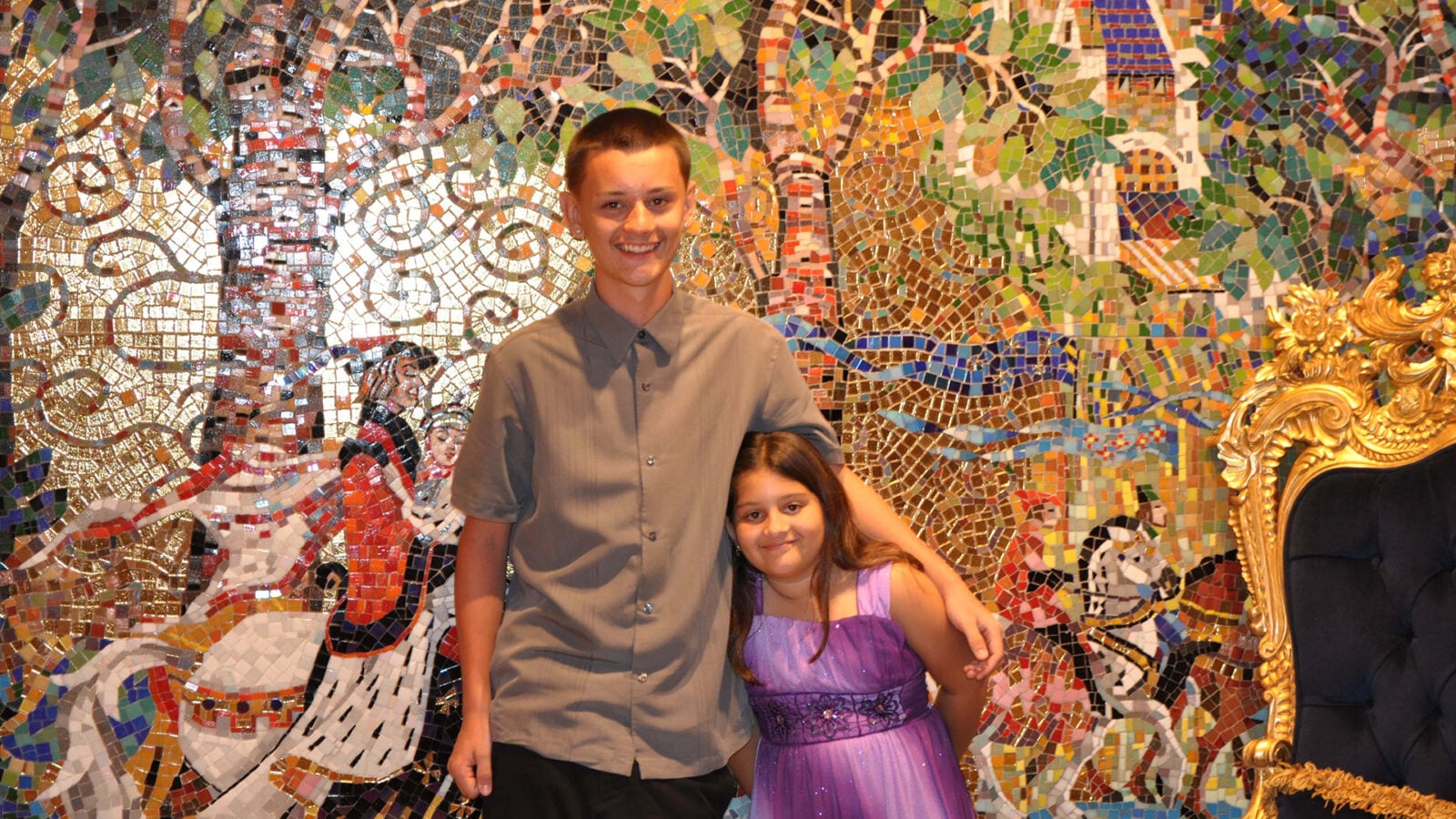Two kids posing in front of a colorful mosaic wall boy in a gray shirt and girl in a purple dress with a gold ornate chair to the right