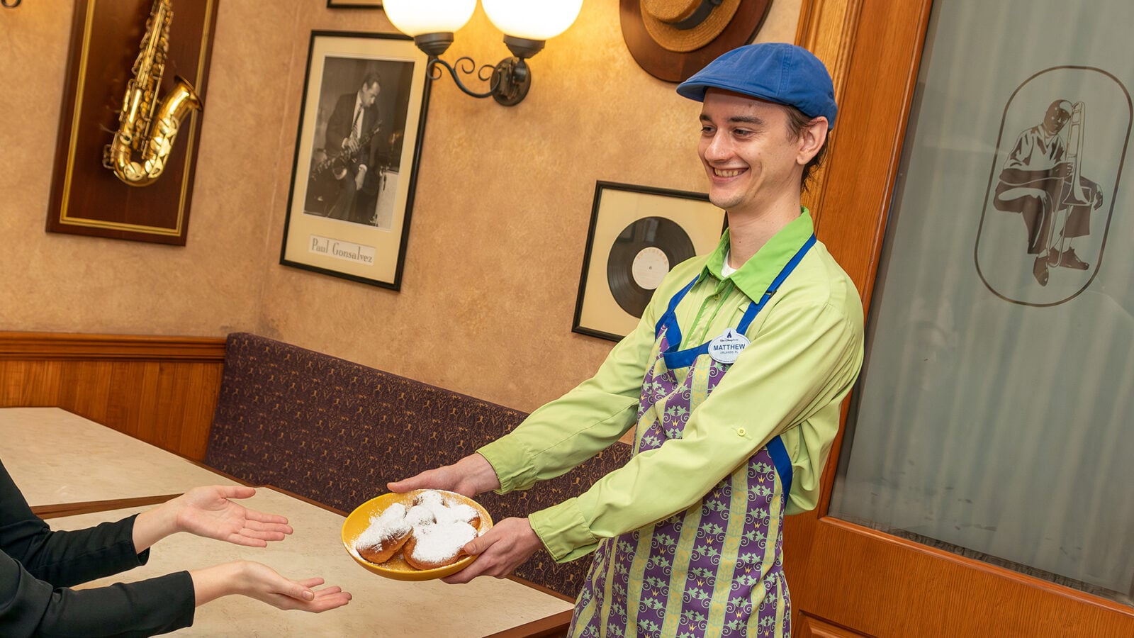 Smiling server in a blue cap and green shirt hands a plate of powdered donuts to a customer in a diner