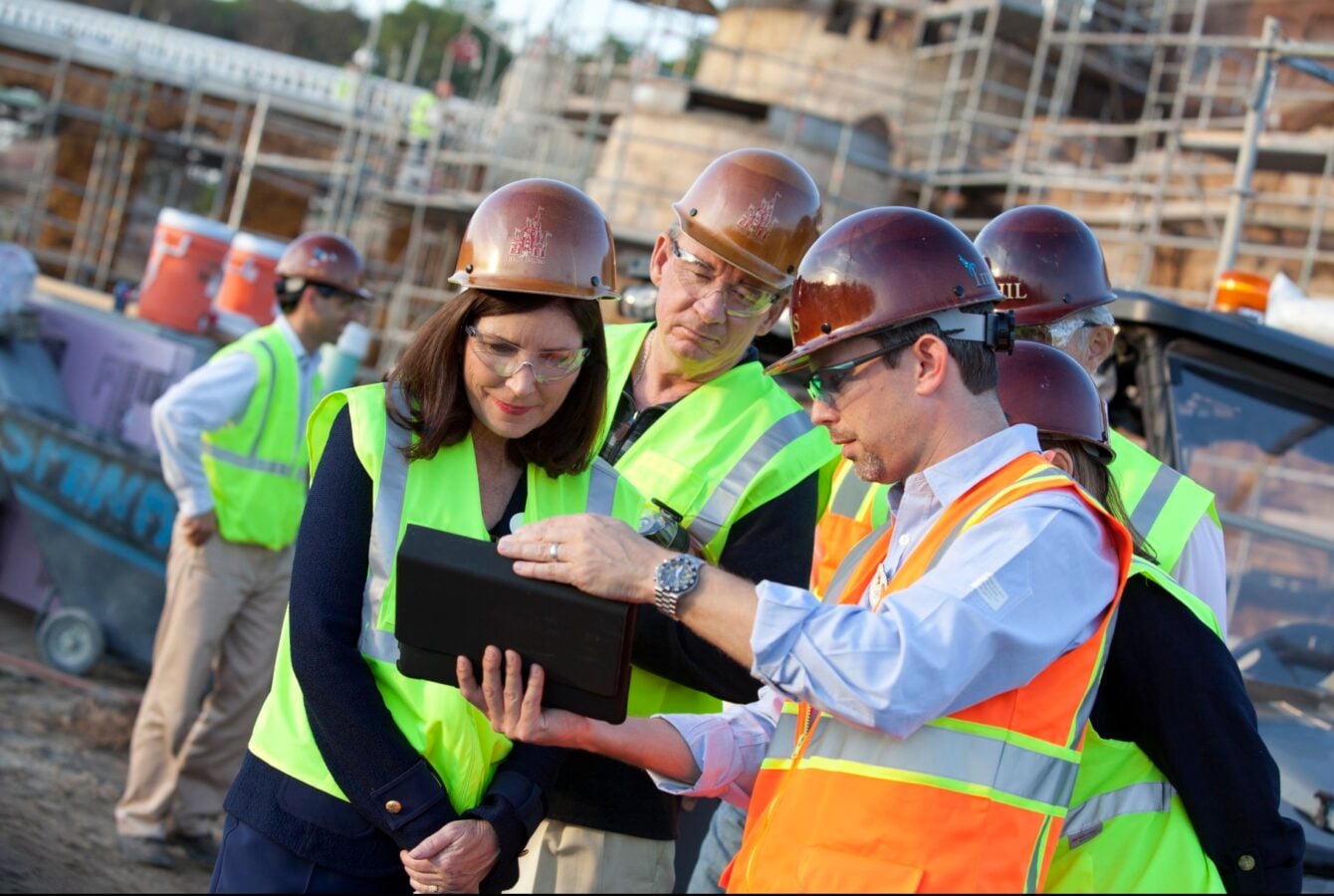 Team of construction workers in hard hats and high visibility vests reviewing a tablet at a busy construction site collaborating closely