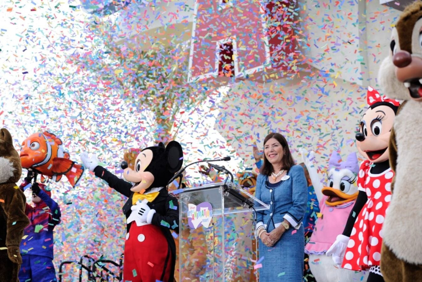 Smiling woman in a blue dress speaks at a podium at a Disney celebration with Mickey Mouse Minnie Mouse and friends as confetti rains down