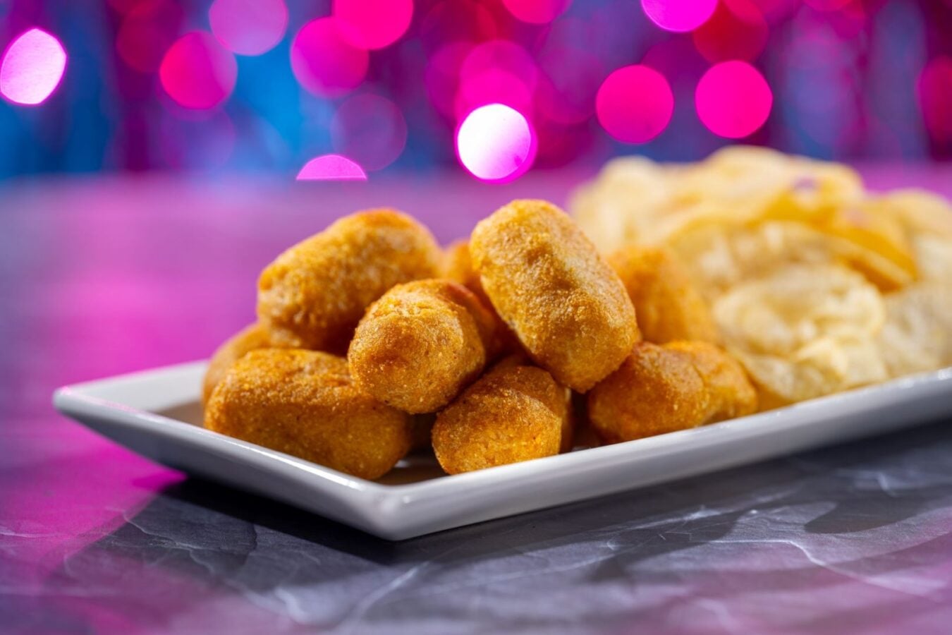 Plate of golden brown croquettes on a white dish with a pile of chips in the background against a pink purple bokeh backdrop