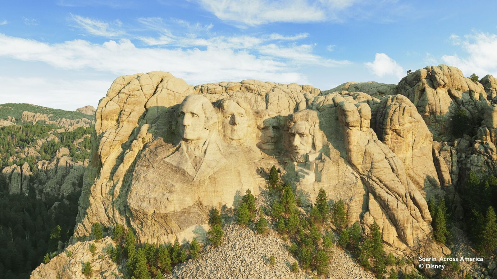 Aerial view of Mount Rushmore with four presidential faces carved into granite set in a rocky mountainside and pine forest beneath a blue sky
