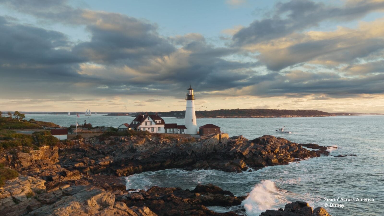 Lighthouse on a rocky coastline with small white houses and boats on calm water at sunset