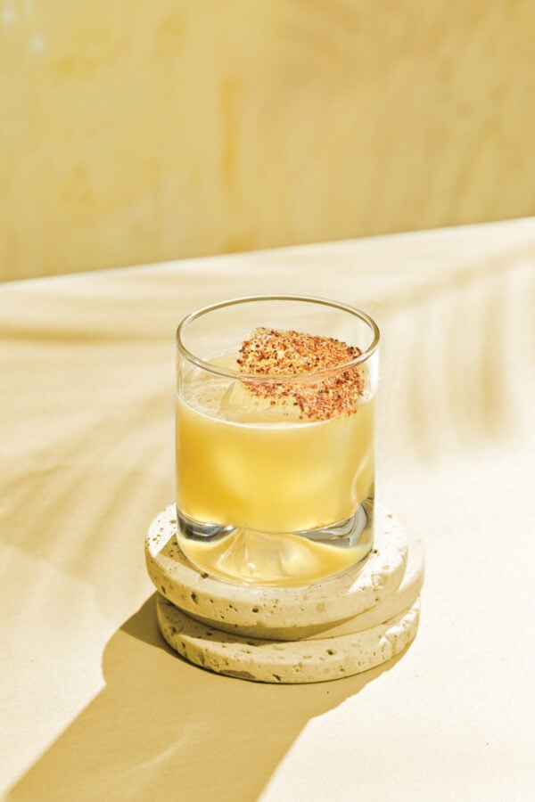 Passionfruit margarita Fantasy Land News Pale yellow cocktail in a short glass with a red chili flake rim set on stacked stone coasters