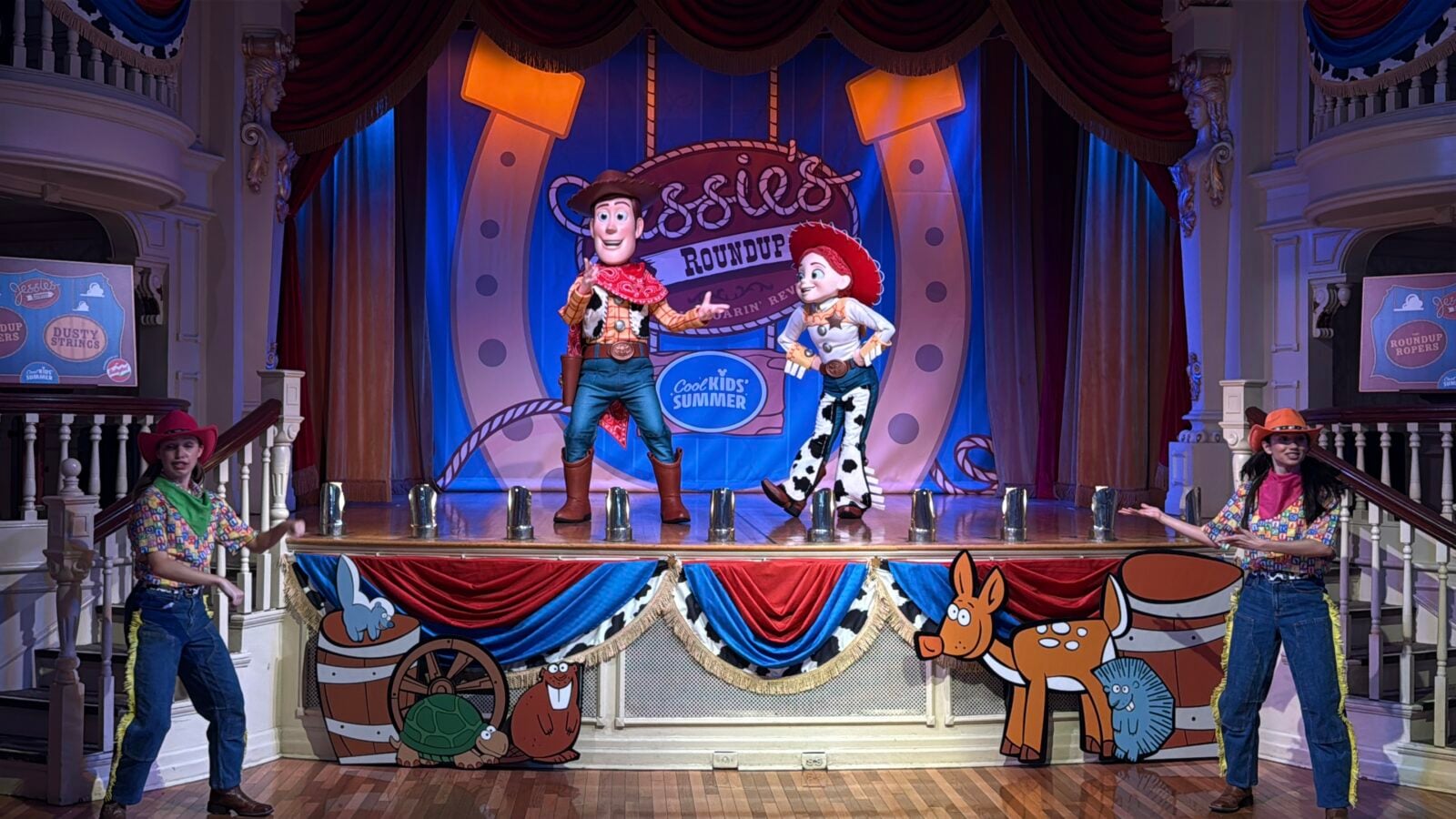 Live stage show with Woody and Jessie characters performing on a colorful western themed set surrounded by cartoon animal props and banners