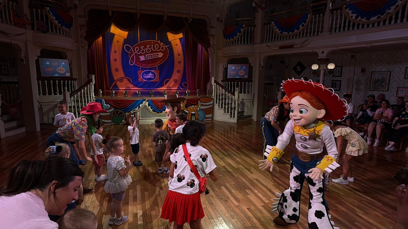 Children interact with life sized Toy Story characters on a theater stage Jessie style cowgirl and a cow patterned character greet kids amid a bright carnival themed backdrop