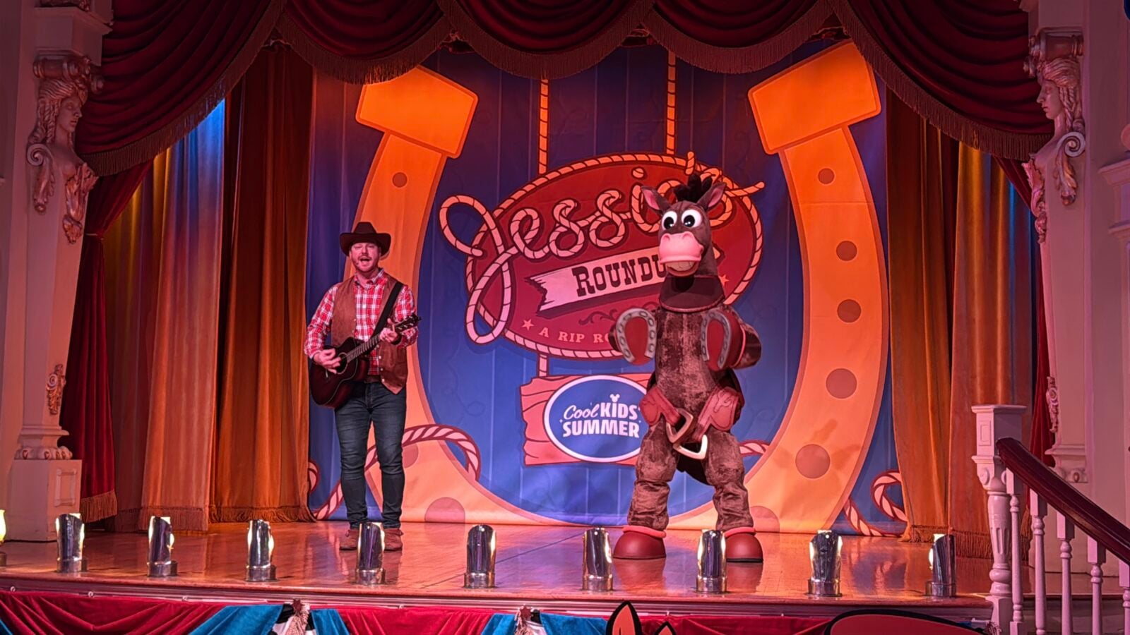 Man in a cowboy hat plays an acoustic guitar on a colorful stage beside a brown horse mascot during a kids show
