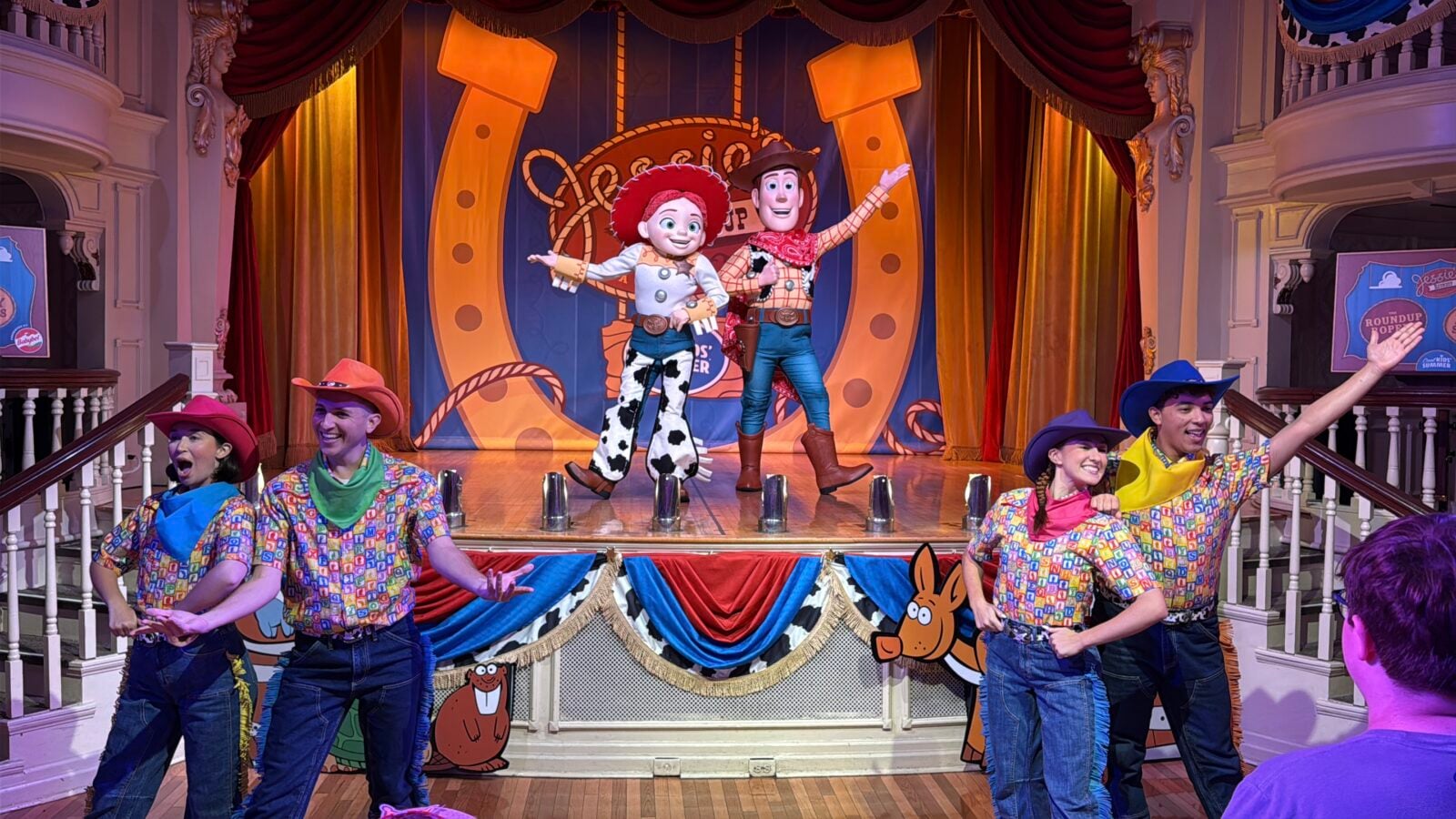 Stage performance featuring Woody and Jessie style puppets center stage with dancers in cowboy hats and colorful shirts in the foreground on a decorated set