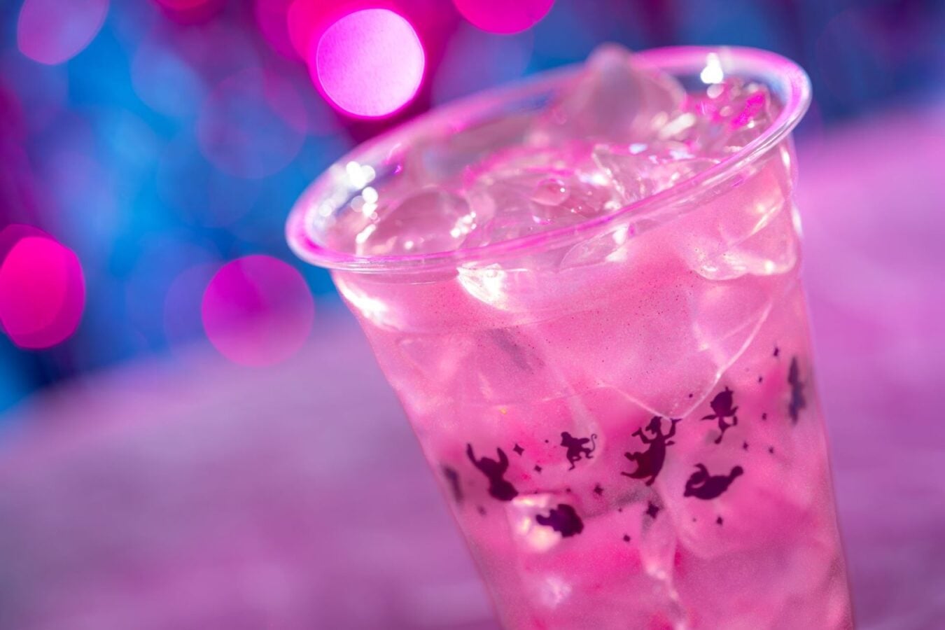 Pink iced drink in a clear plastic cup ice cubes visible cup decorated with fairy silhouettes against a pink purple bokeh background
