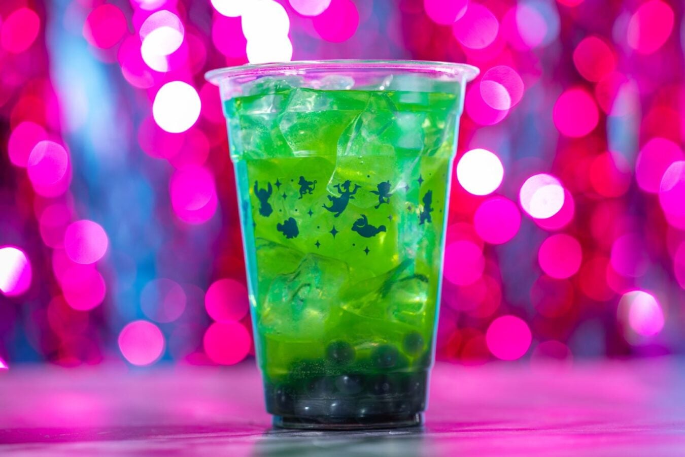 Green iced bubble tea in a clear cup with ice and dark pearls at the bottom against a pink bokeh background