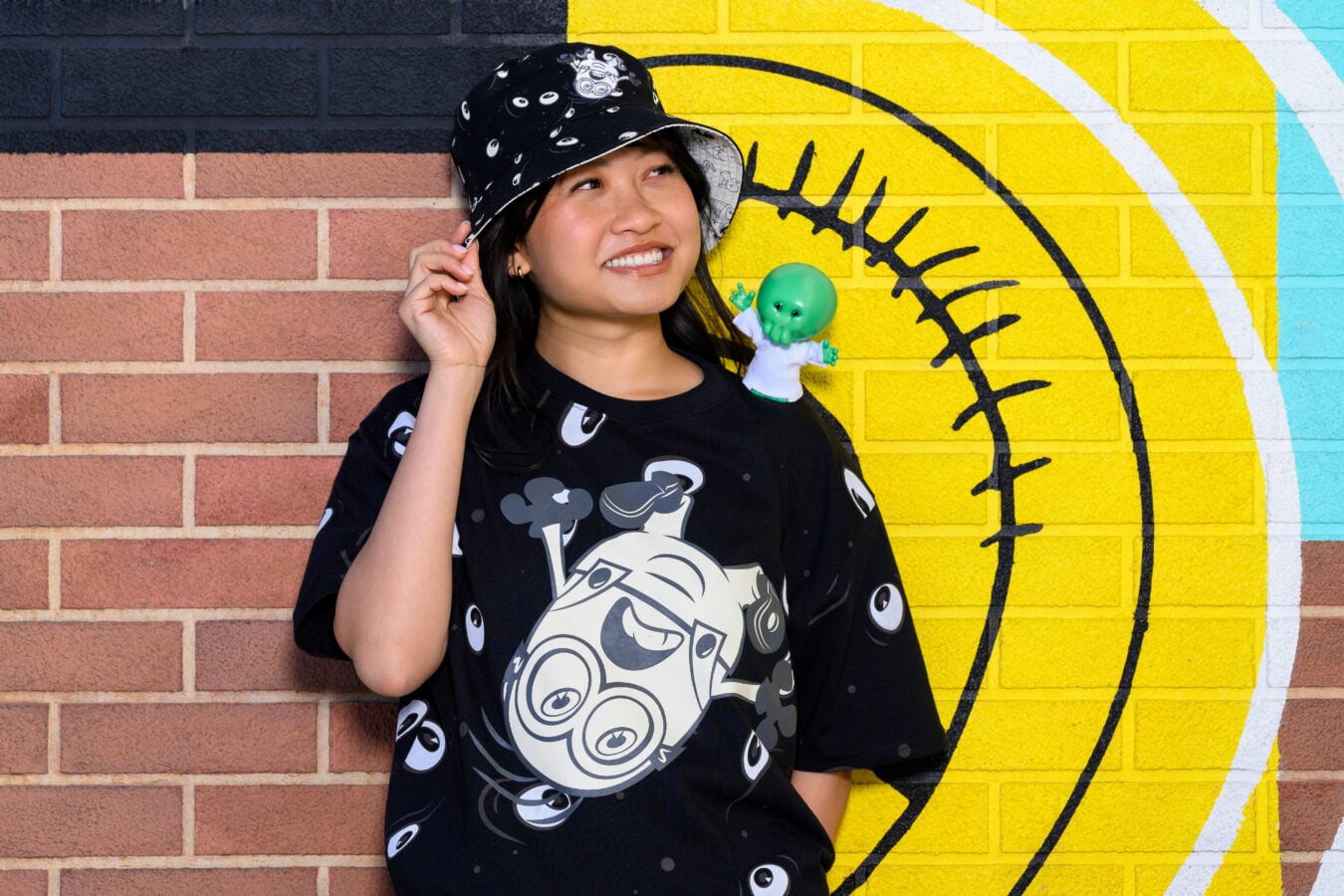 Woman wearing a black graphic tshirt and bucket hat posing against a yellow mural with a green alien toy resting on her shoulderFun character accessory nearby