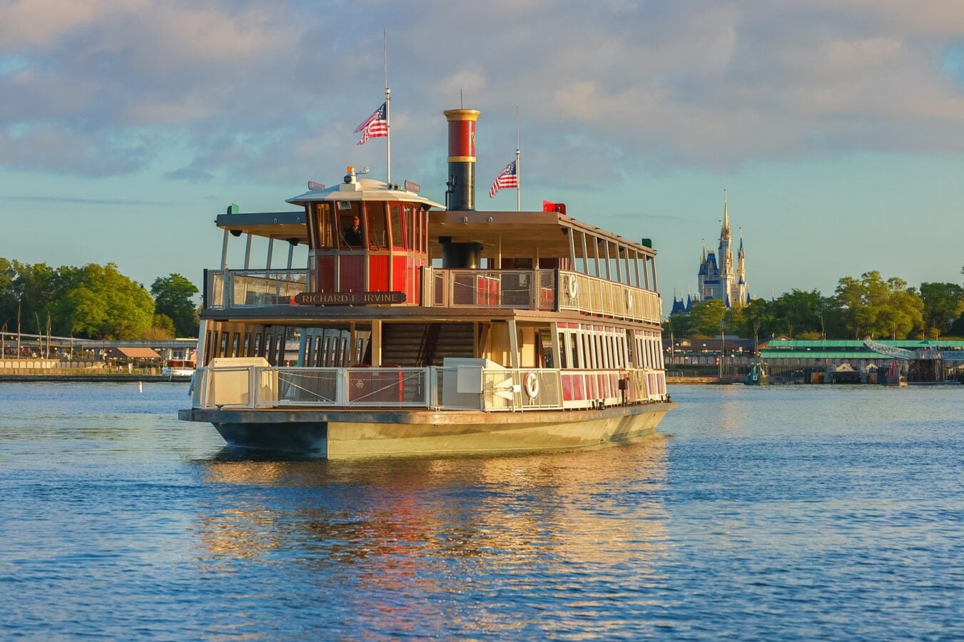 Red and cream passenger riverboat with flags on calm water a distant castle visible on the shore under a blue sky