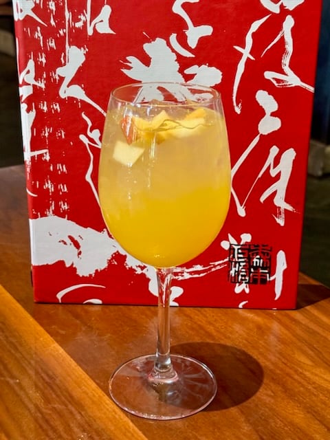 Sake Sangria 2 Fantasy Land News Stemmed wine glass with pale orange cocktail and ice garnished with a citrus twist on a wooden table with a red patterned box in the background