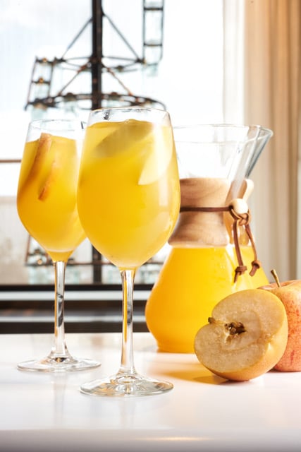 Sake Sangria Fantasy Land News Two glasses of orange juice with ice a pitcher and sliced apples on a white surface by a window