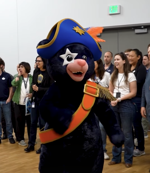 Screenshot 20260518 123802 Fantasy Land News Costumed blue bear mascot in a pirate hat with an orange sash dancing among cheering people indoors