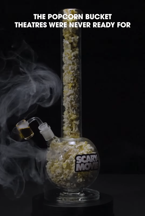 Glass popcorn bucket bong filled with popcorn releasing smoke with a bold white overlay text THE POPCORN BUCKET THEATRES WERE NEVER READY FOR and a SCARY MOVIE label on the base