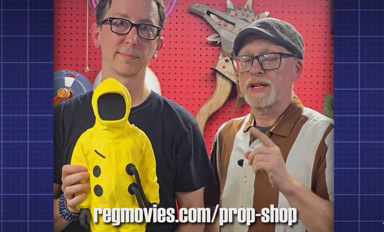 Screenshot 20260521 141233 Fantasy Land News Two men in a workshop hold a bright yellow hooded prop in front of a red pegboard the text regmoviescomprop shop appears at the bottom