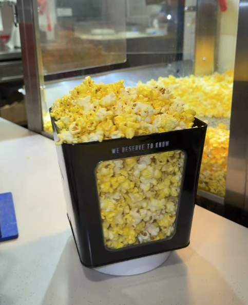 Screenshot 20260527 080756 Fantasy Land News Buttered popcorn overflowing from a black self serve dispenser at a snack counter with a clear window windowed side showing popcorn inside