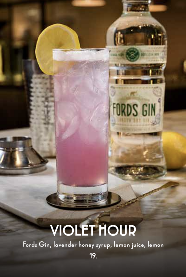 Violet Hour Fantasy Land News A tall glass of pinkish lavender Violet Hour cocktail with a lemon slice beside a Fords Gin bottle on a bar counter menu text reads Violet Hour with ingredients
