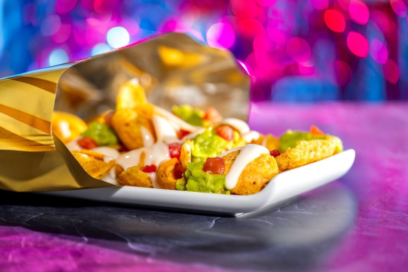 Pile of nachos with guacamole salsa and sour cream on a white plate with a colorful bokeh background