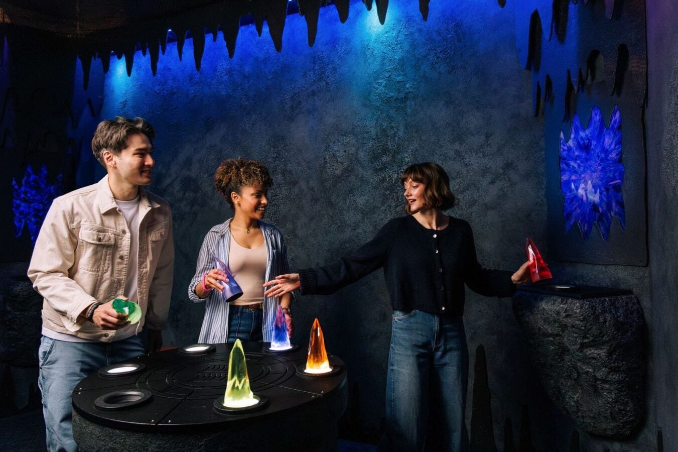 Three friends in a dark blue lit room play with glowing crystal prisms on a circular table sharing and smiling at each other