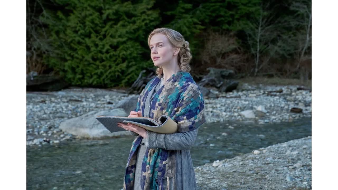 Woman standing by a rocky riverbank holding an open notebook and pen gazing into the distance