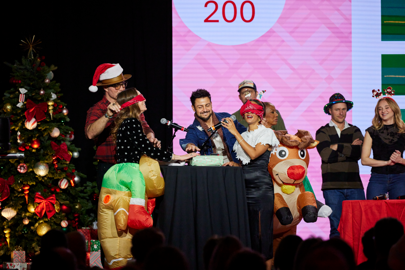 Group of people on a stage at a Christmas event wearing costumes and props with a decorated Christmas tree on the left and gift boxes nearby