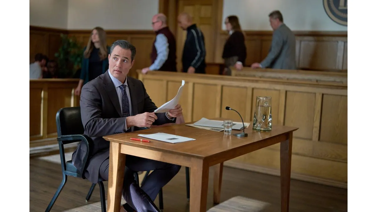 Man in a suit sits at a wooden desk in a courtroom holding papers as a microphone and glass pitcher sit on the desk with people in the background