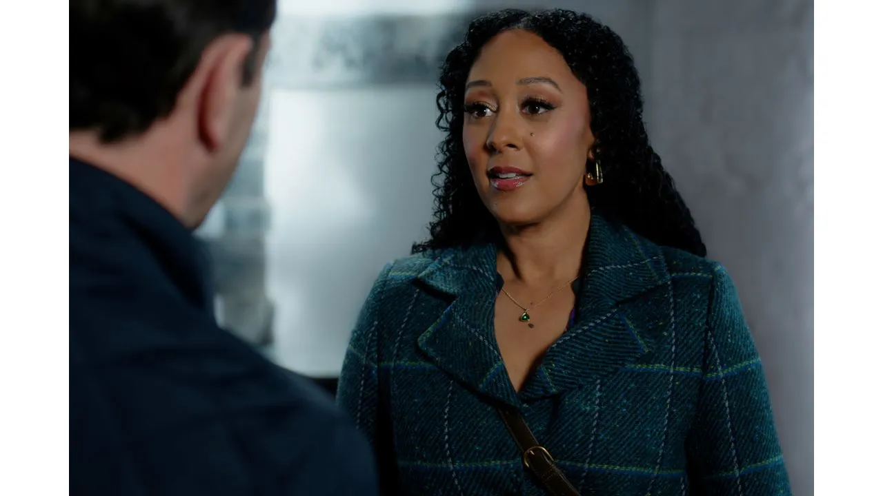 Woman with curly hair wearing a teal plaid coat talks to a man whose back is to the camera in a dim hallway