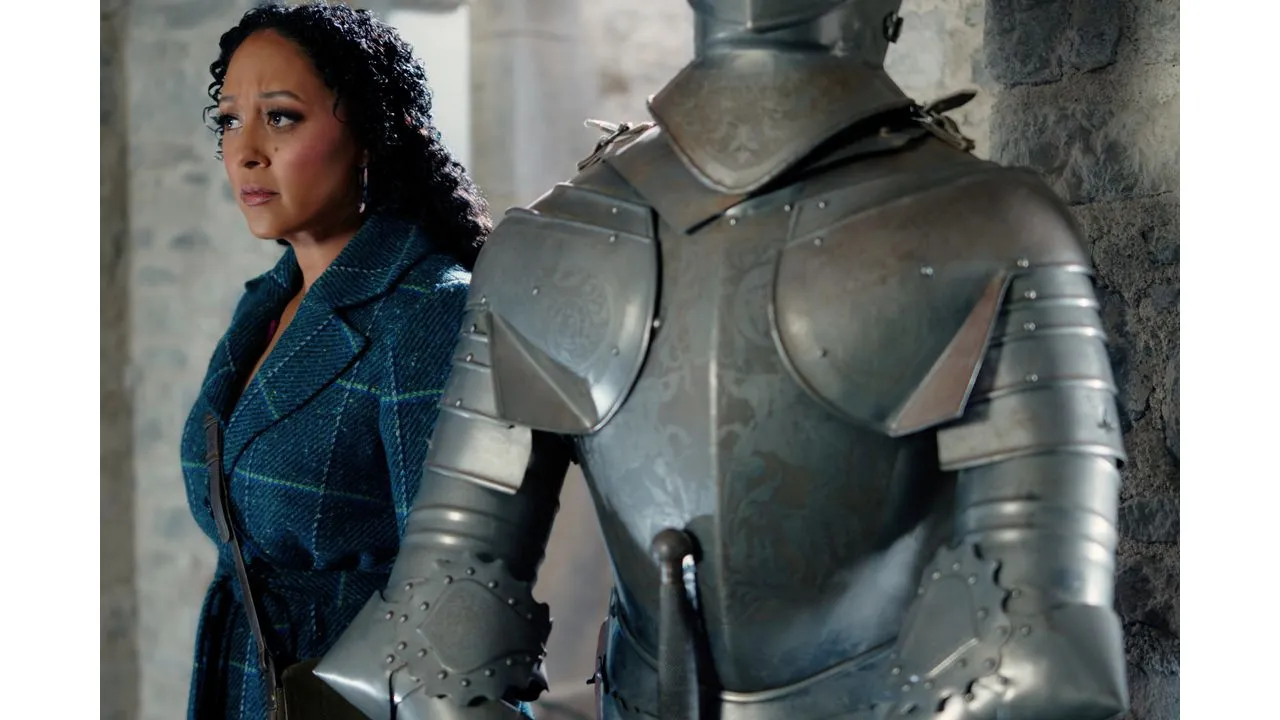 Woman with a blue plaid coat stands beside a suit of armor in a stone walled corridor looking concerned