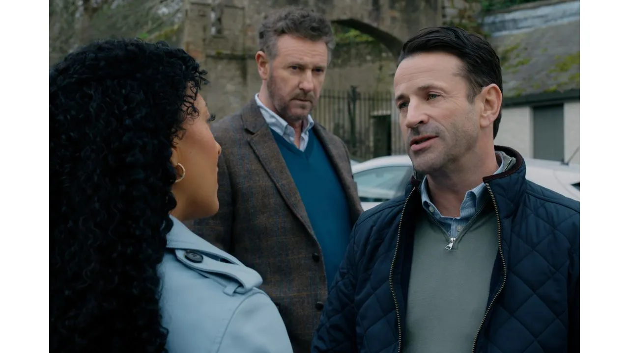 Three adults are having a discussion outdoors with a man in a navy quilted jacket speaking to a woman seen from the side while another man looks on in the background