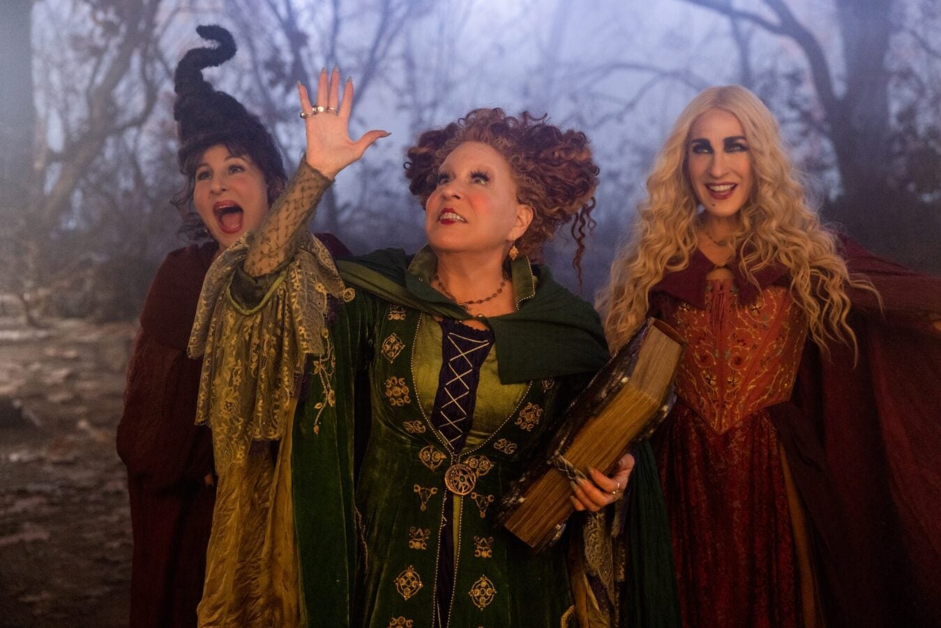 Three women dressed as witches in medieval robes stand in a forest the center figure raising a hand as if casting a spell
