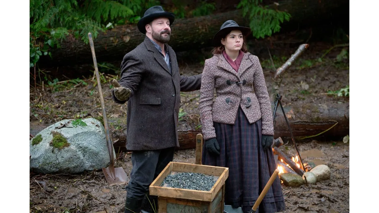 Man and woman in 19th century attire stand by a campfire in a forest with a wooden coal box in front and tools nearby