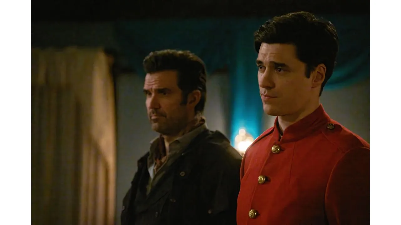 Two men stand in a dim room one in a red military style jacket with gold buttons the other in a dark jacket both looking serious