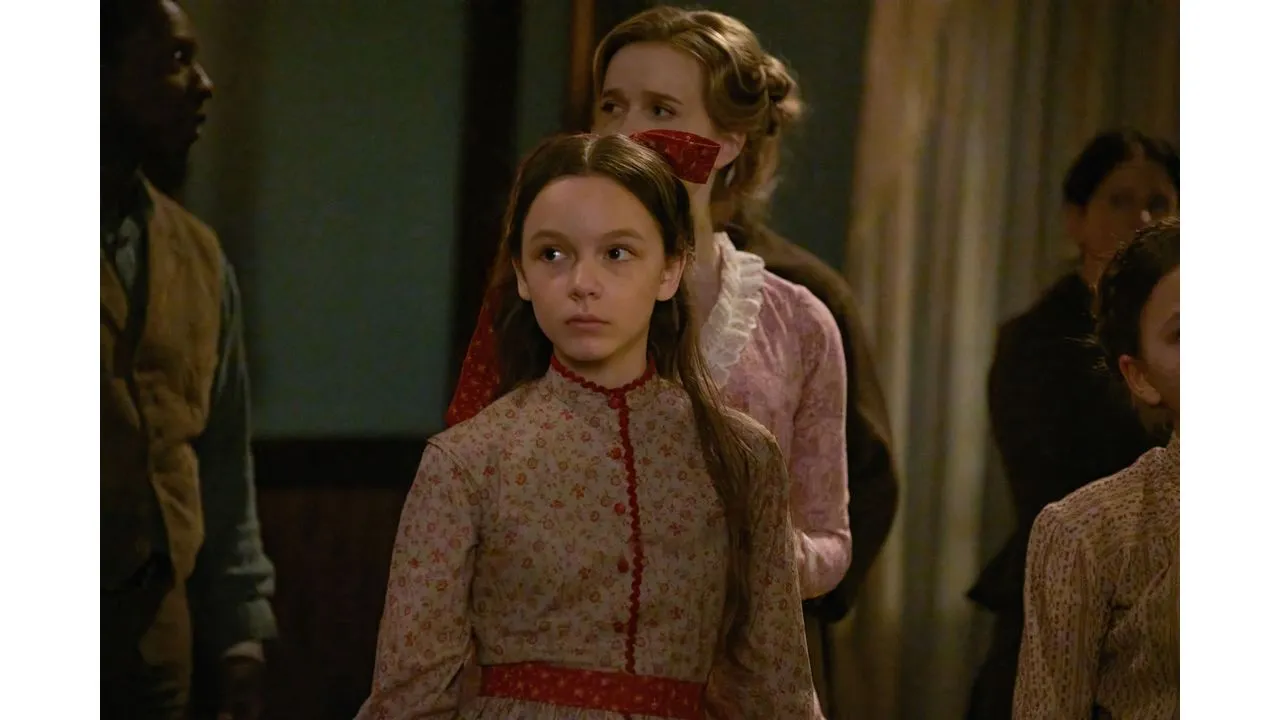 Young girl in a pale floral dress with red trim stands in a dim hallway looking ahead with a serious expression behind her are other people in period clothing