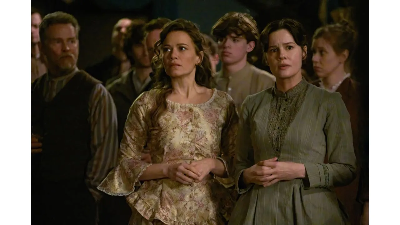 Two women in vintage dresses stand among a crowd looking anxious or concerned in a dimly lit scene The woman on the left wears a floral brocade dress the woman on the right wears a pale green striped dress