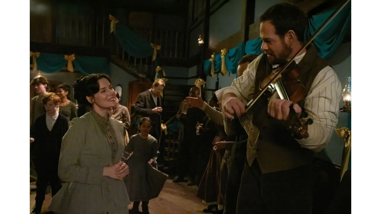 Woman in a vintage green dress smiles while a man plays the violin at a crowded retro looking party in a decorated hall